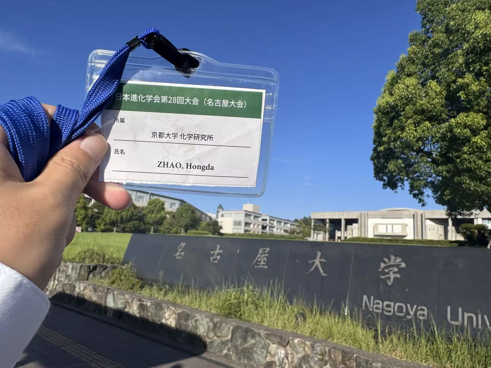 A conference badge bearing Hongda Zhao’s name is held up in front of the Nagoya University entrance sign beneath a clear blue sky.